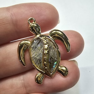 Puede incluir: Un colgante de tortuga de color dorado con una concha nacarada. El colgante tiene un lazo en la parte superior para colgarlo y probablemente se utiliza para la fabricación de joyas. La concha de la tortuga tiene una calidad brillante e iridiscente.