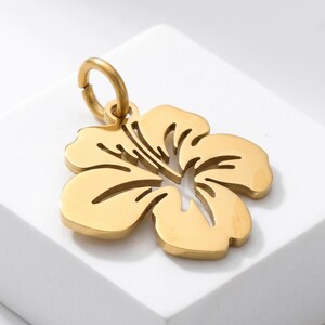 Puede incluir: Colgante dorado con forma de flor de hibisco. El colgante tiene un dise&ntilde;o calado y un peque&ntilde;o lazo en la parte superior para sujetarlo a una cadena o pulsera. La flor tiene cinco p&eacute;talos y l&iacute;neas interiores detalladas.