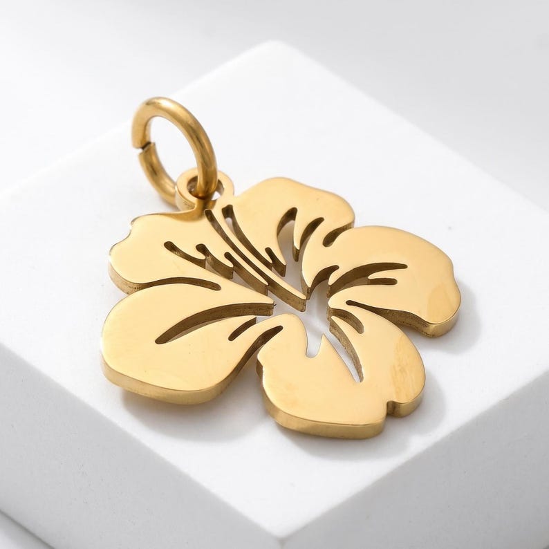Puede incluir: Colgante dorado con forma de flor de hibisco. El colgante tiene un dise&ntilde;o calado y un peque&ntilde;o lazo en la parte superior para sujetarlo a una cadena o pulsera. La flor tiene cinco p&eacute;talos y l&iacute;neas interiores detalladas.