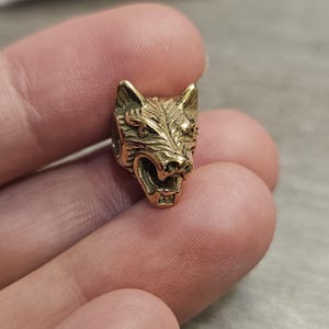 May include: A small, metallic gold-toned bead shaped like a snarling wolf head. The wolf head features detailed fur textures, pointed ears, and an open mouth showing teeth. The surface has a polished, reflective finish with dark recessed areas.
