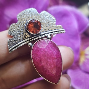 May include: A silver-toned ring featuring a large, teardrop-shaped, magenta-colored gemstone and a smaller, round, orange-red gemstone. The ring has a textured, floral-inspired design around the smaller stone.