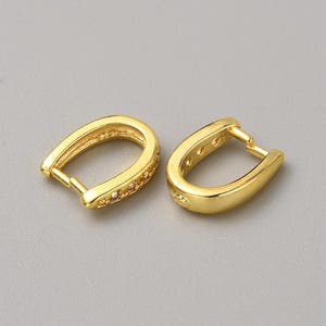May include: Two gold-colored hoop earrings. One earring has small, clear stones set along the outer curve. The other earring is a solid gold color. Both earrings have a hinged closure and are set against a gray background.