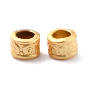 May include: Two gold-colored cylindrical beads with a matte finish. Each bead features a central hole and is adorned with an embossed, swirling pattern around its circumference. The beads are displayed against a white background.