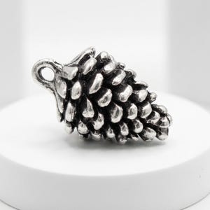 Op de afbeelding: Een zilverkleurige dennenappelhanger met een getextureerd oppervlak. De hanger heeft een lus aan de bovenkant om aan een ketting of koord te bevestigen. De dennenappel is gedetailleerd met individuele schubben.