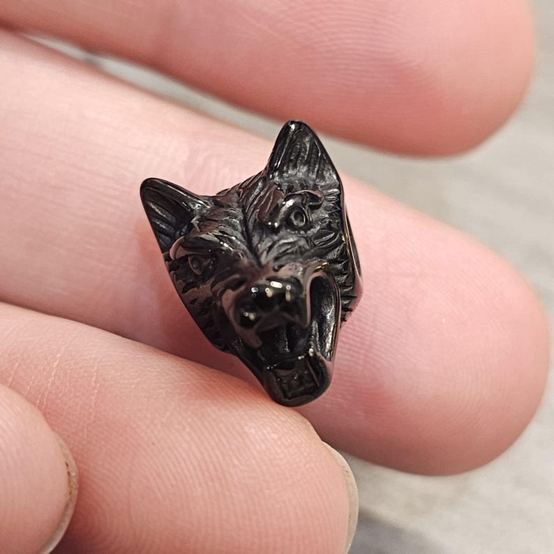 May include: A small, dark metallic bead shaped like a snarling wolf head. The charm features detailed fur textures, pointed ears, and an open mouth with visible teeth, designed with a polished finish that reflects light across its sculpted surface.
