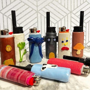 Pode incluir: Uma coleção de isqueiros decorativos com vários designs. Os isqueiros apresentam enfeites únicos, incluindo um laço azul, um cogumelo e um rosto de desenho animado. Os isqueiros são em tons de vermelho, azul, castanho e branco.