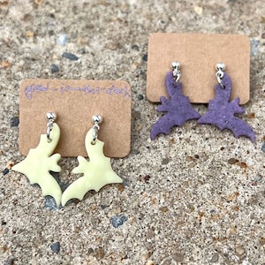 Puede incluir: Dos pares de pendientes en forma de murciélago. Un par es de color amarillo claro y el otro par es de color morado claro. Los pendientes están en una tarjeta marrón con el texto "glow in the dark" escrito en ella.