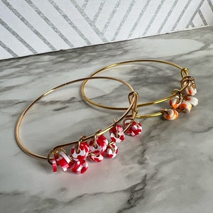 May include: Two gold-colored bangles with red and white candy-shaped charms. The charms are round with a swirl pattern. The bangles are displayed on a marble surface with a white and silver geometric patterned background.