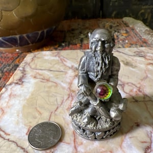May include: A pewter figurine of a dwarf holding a pickaxe and a gem. The dwarf is wearing a hat and has a beard. The gem is iridescent and has a red, green, and yellow color.