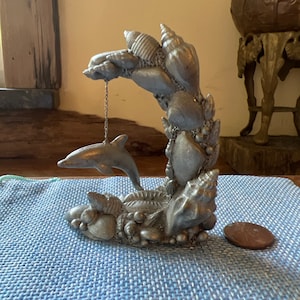 以下が含まれることがあります： 鎖で吊るされたイルカの銀色の彫刻で、背景には貝殻があります。ベースにはさらに貝殻と小さな岩があります。彫刻の隣にはペニーがあります。彫刻は青い表面にあります。