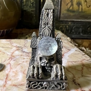 May include: A silver metal figurine of a skull with a tower on top. The skull is holding a crystal ball in its hands. The tower is detailed with brickwork and a pointed roof. The figurine is sitting on a base with a snake design.