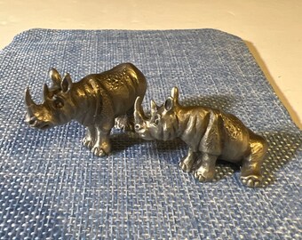 May include: Two pewter rhinoceros figurines, one facing left and the other facing right, on a blue textured surface.