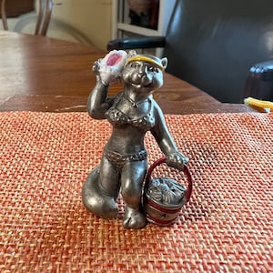May include: A pewter figurine of a cat wearing a bikini and a headband, holding a seashell and a bucket with seashells inside.