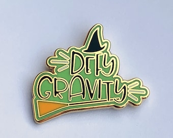 May include: A green enamel pin with the words "Dirty Gravity" in white lettering. The pin features a witch's hat, a broom, and gold-colored trim. The pin is on a white background.