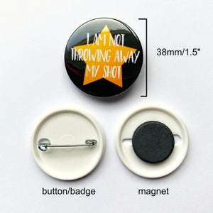 May include: A black and gold button badge with the text "I AM NOT THROWING AWAY MY SHOT" in white lettering. The badge is 1.5 inches in diameter and has a safety pin on the back.