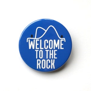 May include: Blue round button with white text that says "Welcome to the Rock" and a white outline of a mountain range with two small trees on either side.