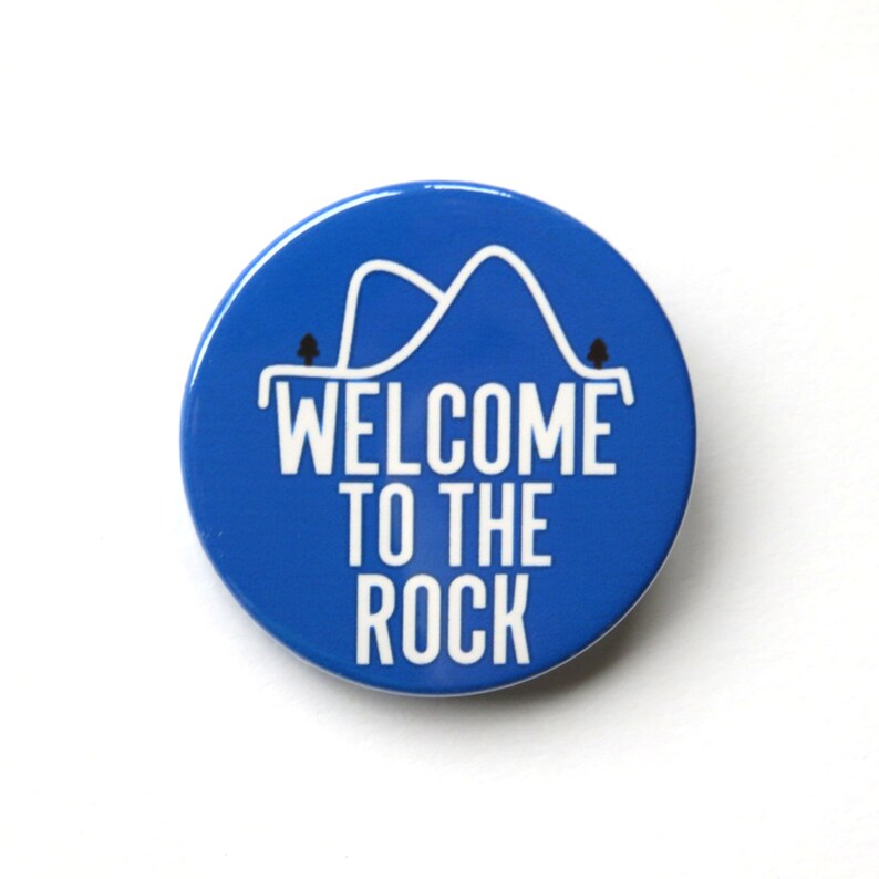 May include: Blue round button with white text that says "Welcome to the Rock" and a white outline of a mountain range with two small trees on either side.