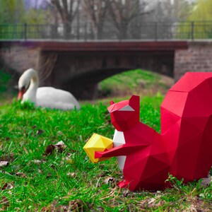 May include: A red origami squirrel holding a yellow origami nut. The squirrel is sitting on green grass in front of a bridge.