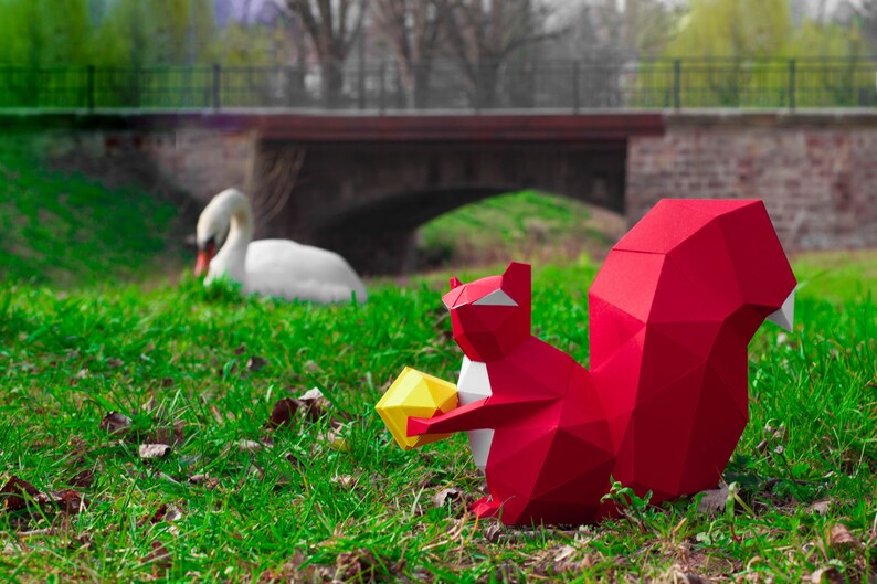 May include: A red origami squirrel holding a yellow origami nut. The squirrel is sitting on green grass in front of a bridge.