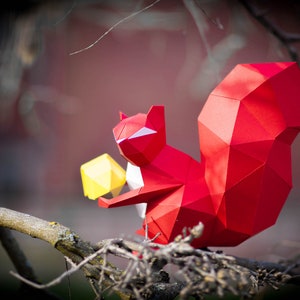 May include: A red origami squirrel holding a yellow origami nut. The squirrel is perched on a branch with a blurred background.