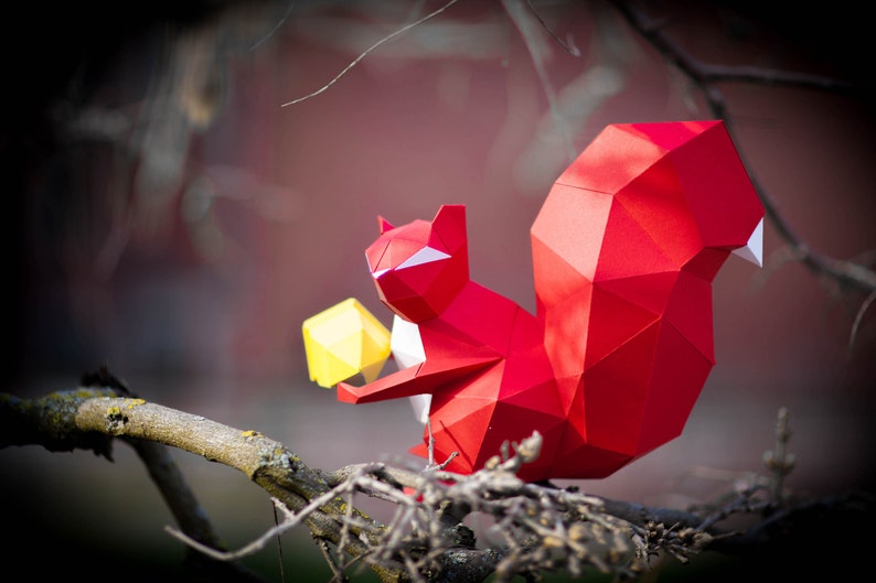 May include: A red origami squirrel holding a yellow origami nut. The squirrel is perched on a branch with a blurred background.