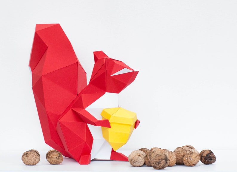 May include: A red origami squirrel holding a yellow origami nut. The squirrel is standing on a white surface with several brown walnuts scattered around it.