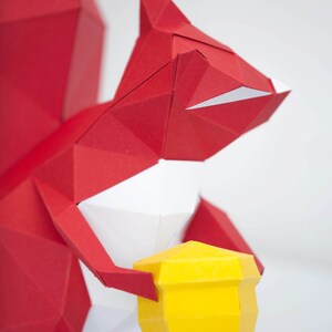 May include: A red origami squirrel holding a yellow origami acorn. The squirrel is made of paper and has a white belly.