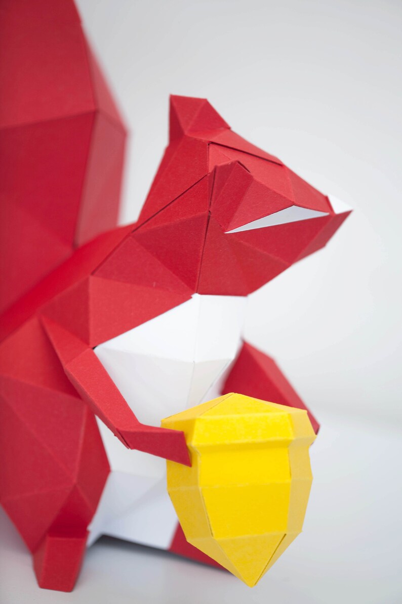 May include: A red origami squirrel holding a yellow origami acorn. The squirrel is made of paper and has a white belly.