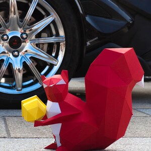 May include: A red origami squirrel holding a yellow origami nut. The squirrel is sitting on a gray concrete surface in front of a black car.