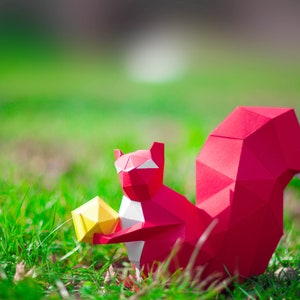 May include: A red origami squirrel holding a yellow origami nut in its paws. The squirrel is sitting on a bed of green grass.