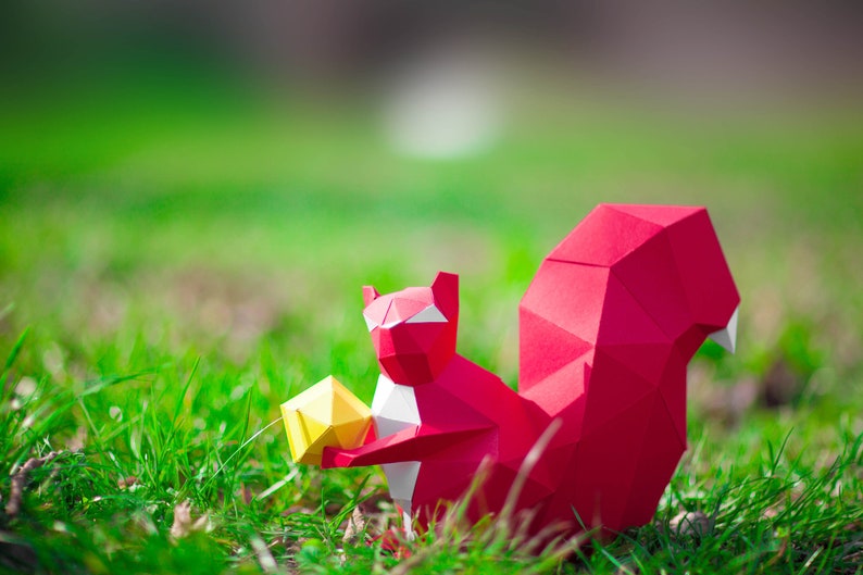 May include: A red origami squirrel holding a yellow origami nut in its paws. The squirrel is sitting on a bed of green grass.