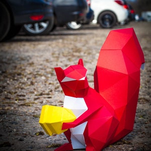 May include: A red origami squirrel holding a yellow origami nut. The squirrel is standing on a gravel surface with cars in the background.