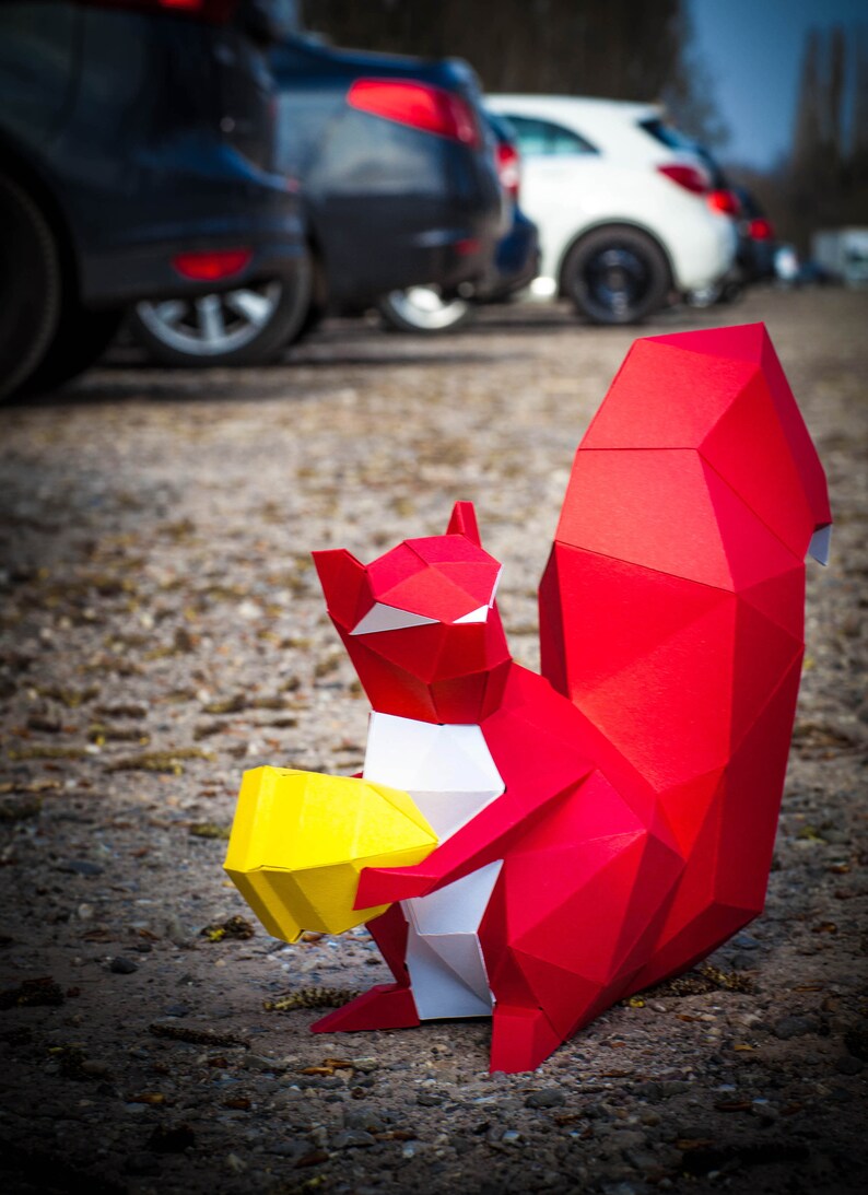 May include: A red origami squirrel holding a yellow origami nut. The squirrel is standing on a gravel surface with cars in the background.