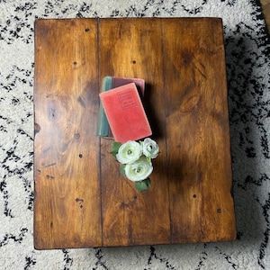May include: A dark brown wooden coffee table with a natural wood grain finish. The table has a square top and is decorated with a stack of books and white flowers.