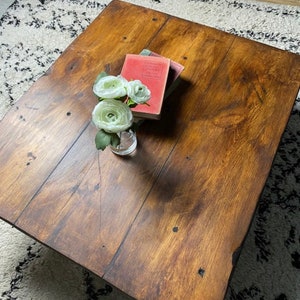 May include: A rustic wooden coffee table with a natural wood finish. The table is rectangular and made of thick planks of wood. The table is decorated with a small bouquet of white flowers in a clear glass vase. There are three books stacked on top of the table, with the top book titled "The Complete Works of William Shakespeare".