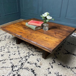 May include: A rustic wooden coffee table with a natural wood finish and a rectangular shape. The table has a simple design with two thick wooden legs on each side. The table is sitting on a white and black patterned rug.