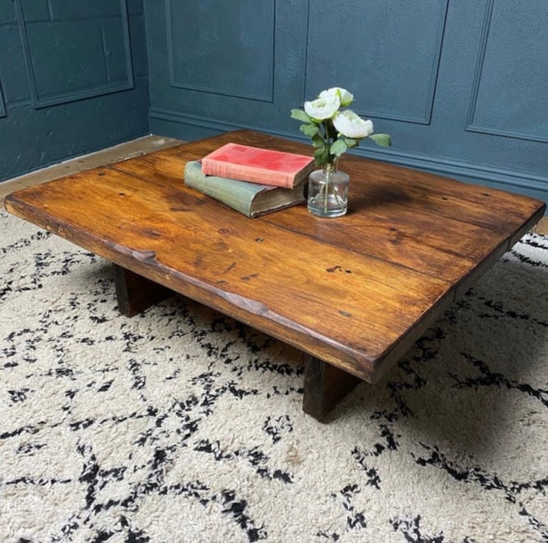 May include: A rustic wooden coffee table with a natural wood finish and a rectangular shape. The table has a simple design with two thick wooden legs on each side. The table is sitting on a white and black patterned rug.