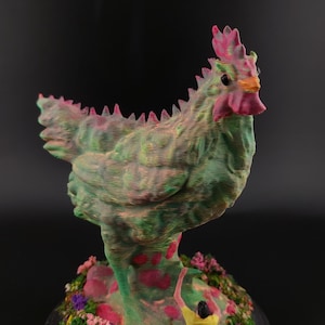 Puede incluir: Una escultura caprichosa de una criatura parecida a un pollo con una combinación de colores verde y rosa. La escultura texturizada está colocada sobre una base con flores coloridas y una pequeña figura. El diseño general es artístico y único.