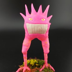 Könnte beinhalten: Rosa Monsterfigur mit breitem Grinsen und stacheligem Kopf. Die Kreatur hat gelbe Augen und steht auf einem Sockel aus Moos und künstlichem Laub. Die Figur ist etwa 10 cm hoch.