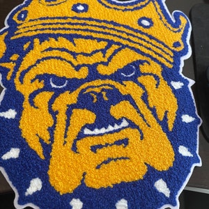 Op de afbeelding: Een geborduurde patch met een bulldog die een kroon draagt. De bulldog is geel en blauw, heeft een woeste uitdrukking en is omringd door een witte rand.