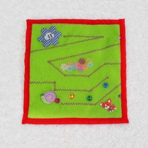 May include: A green felt board with a red border. The board has a variety of felt shapes and buttons attached to it, including a flower, a snail, a ladybug, and a fox. The board is designed for children to play with and learn about different shapes and colors.