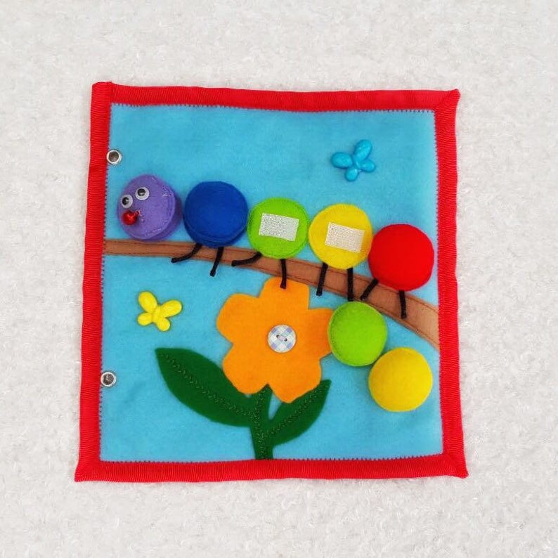 May include: A felt book for toddlers featuring a colorful caterpillar on a branch with a flower and a butterfly. The caterpillar has a purple head, blue, green, yellow, and red segments. The flower is orange with a green stem and leaves. The butterfly is blue.