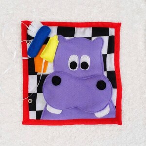May include: A purple felt hippopotamus with black eyes and a black nose is attached to a black and white checkered fabric square. The fabric square is surrounded by a red border. There are felt objects attached to the top of the fabric square, including a blue toothbrush, a yellow tube of toothpaste, and an orange toothbrush.
