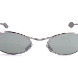 May include: Silver-framed sunglasses with oval lenses and a subtle green tint. The Ray-Ban logo is visible on the left lens. The temples have gray ear pieces. A stylish accessory for eye protection.