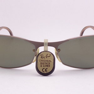Puede incluir: Un par de gafas de sol Ray-Ban plateadas con lentes marrones. Las gafas tienen una etiqueta adjunta que dice "Original Bausch & Lomb CE".