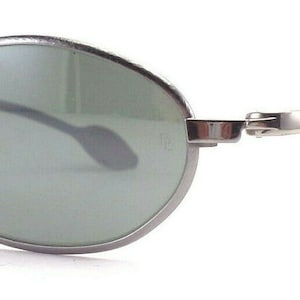 May include: Oval-shaped sunglasses with a silver metal frame and dark green lenses. The sunglasses have a sleek, modern design with a curved bridge and temples. The nose pads are gray. The overall look is minimalist and stylish.