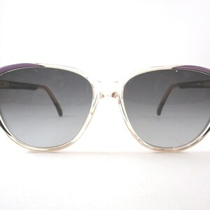 Könnte beinhalten: Transparente Cat-Eye-Sonnenbrille aus Kunststoff mit violettem Rand und grauen Gläsern.