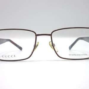 May include: Burgundy-colored metal eyeglasses with clear lenses. The frames are rectangular and have the word "GUCCI" printed on the left side and "STAINLESS STEEL" on the right side.