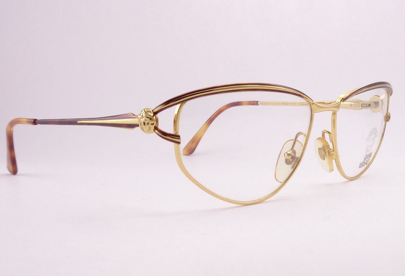 May include: Gold-tone cat-eye glasses with brown tortoise shell arms and a decorative detail on the bridge. The glasses have clear lenses.