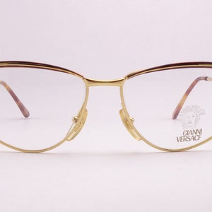May include: Gold-tone metal eyeglasses with a brown accent on the top of the frame. The glasses have a cat-eye shape and the brand name "GIANNI VERSACE" is printed on the right lens.